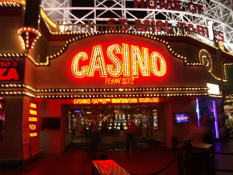Night photo of the casino entrance: giant red channel letters filled with bulbs spelling CASINO, Penny Slots in neon script below, HOME OF $3.00 MINIMUM TABLES in gold neon over the doors, and the lit white coaster structure above.