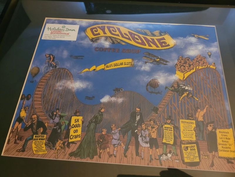 A framed blue and yellow Cyclone Coffee Shop print photographed behind glass: a Victorian wooden roller coaster crowded with cartoon riders, biplanes and balloons in the sky, joke classified signs on the boardwalk, and a Holiday Inn Casino Boardwalk address sticker at top left.