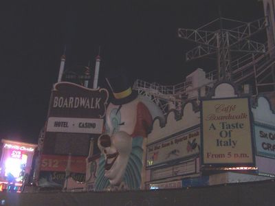 Night photo of the closed Boardwalk: the giant clown face and BOARDWALK HOTEL CASINO marquee completely unlit against a black sky, with only a Caffe Boardwalk menu sign and neighboring Monte Carlo lights still glowing.