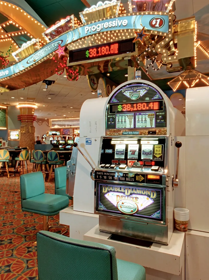A dollar Double Diamond Deluxe slot machine beneath the Boardwalk’s bulb-lit Progressive arch, its jackpot meter reading 38,180 dollars, with teal chairs and the table games behind it.