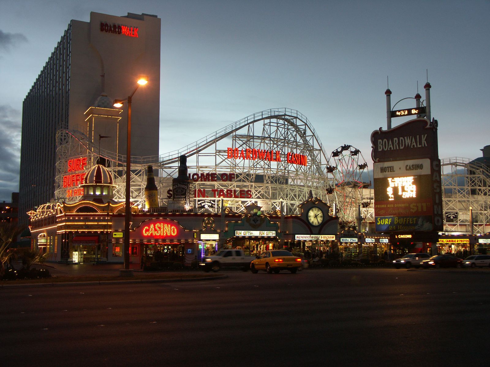 The full Boardwalk property at dusk: white tower with red BOARDWALK letters, lit roller coaster facade, Ferris wheel, glowing casino entrance and the tall price-card pylon, with street traffic in front.