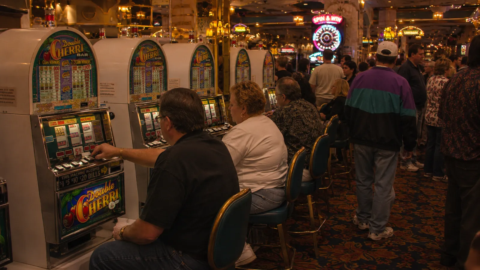 A crowded Boardwalk slot aisle: players seated at Double Cherri Bar machines while a crowd moves down the patterned carpet behind them, neon glowing at the far end.