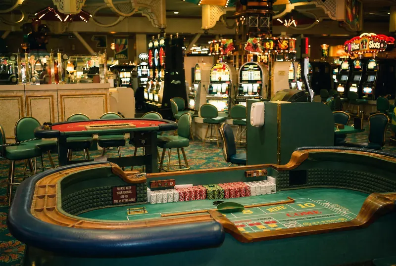 An empty Boardwalk craps table and nearby card table surrounded by green casino chairs, with rows of glowing slot machines beyond.