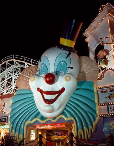 Giant clown face at night: white fiberglass face with pale blue ruffled collar, tan curls, black top hat and a dark red grin, mounted above a casino entrance with coaster framework behind.
