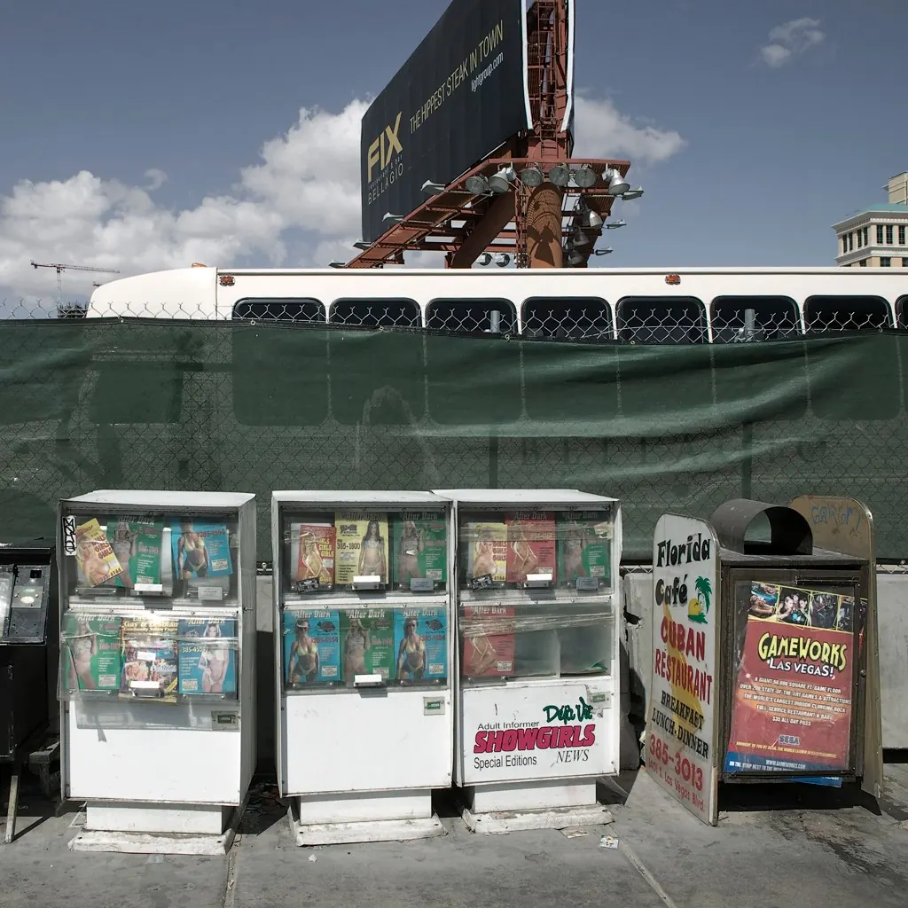 Las Vegas Boulevard sidewalk outside the Boardwalk area with newspaper boxes, restaurant signs, a bus, a billboard structure and construction fencing.