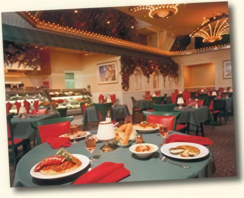 A dining room with hunter green tablecloths, red napkins and chairs, crystal chandeliers, rose garlands on the walls and plates of food in the foreground.