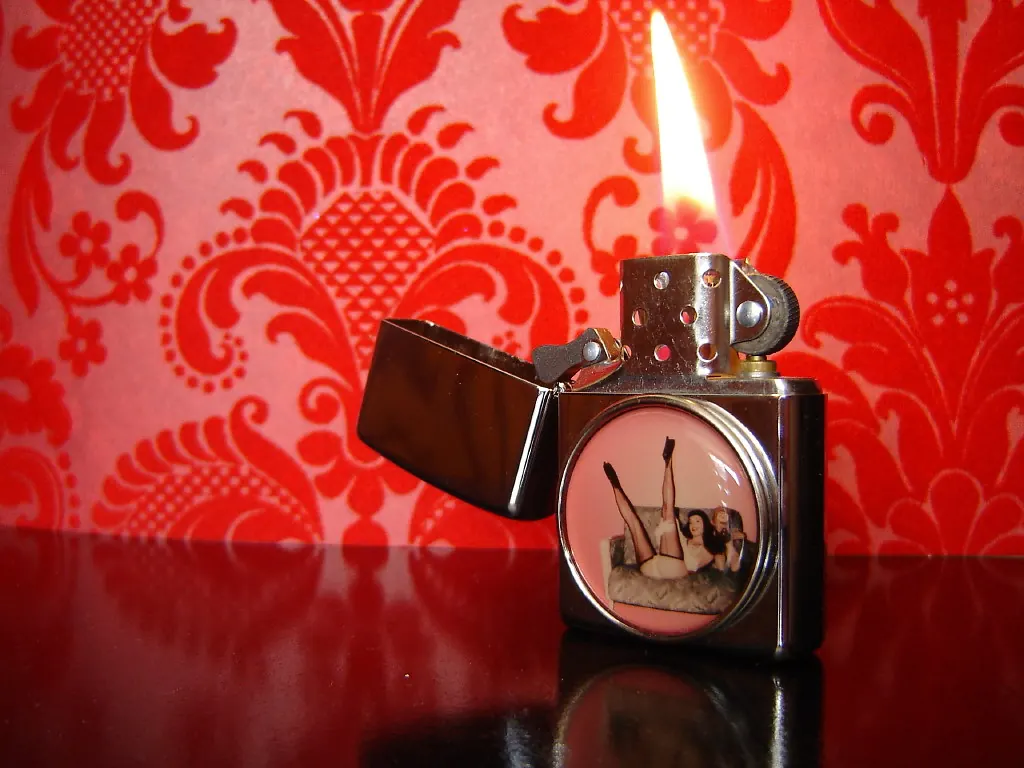 Period novelty lighter printed with a pin-up picture, photographed with its flame lit.