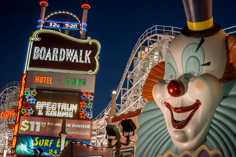 Night photo looking up at the Boardwalk pylon sign: a lit reader board listing Spectrum and an $11.99 prime rib dinner framed by multicolored neon stars, with the giant clown head and white coaster lattice against the dark sky at right.