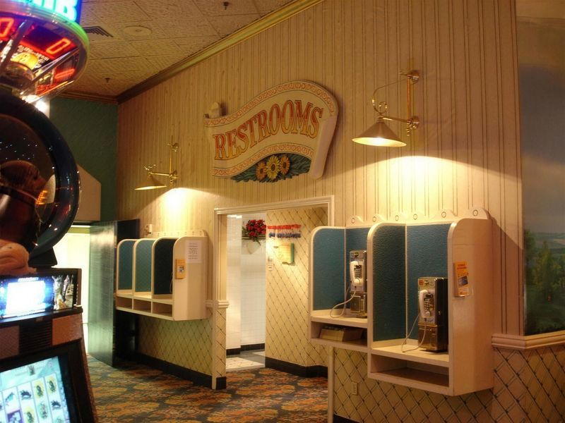 Casino restroom entrance with cream beadboard walls: a hand-painted scroll sign reading RESTROOMS decorated with sunflowers, two brass swing-arm lamps, and white payphone carrels with blue tweed panels holding chrome payphones.