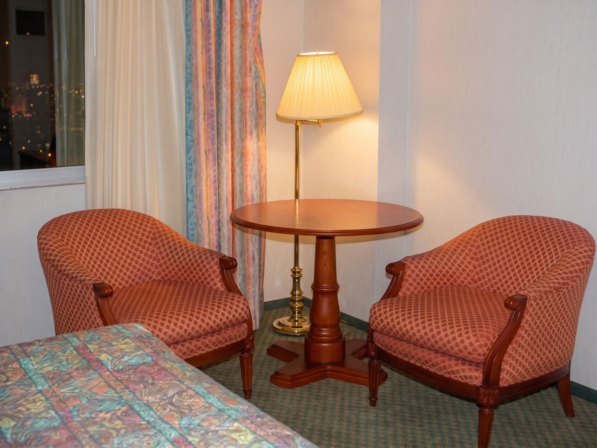 A round wood table and two rose-patterned upholstered chairs beside a brass floor lamp, with a half-drawn curtain and the lit city visible through the tower window.