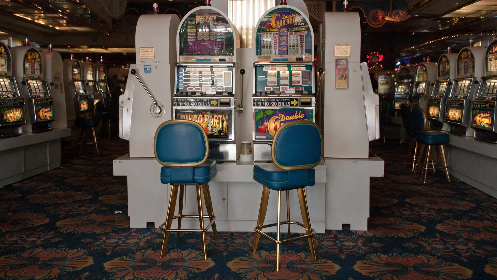 Two white Boardwalk slot machines with blue-and-gold stools and a paper cup between them, centred in a wide view down the patterned-carpet slot aisle.