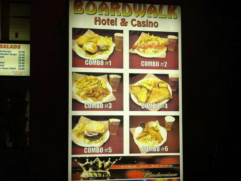 Backlit menu board reading BOARDWALK Hotel & Casino with photos of six numbered food combos over a Budweiser King of Beers panel, beside a smaller lit board listing salads and burgers.