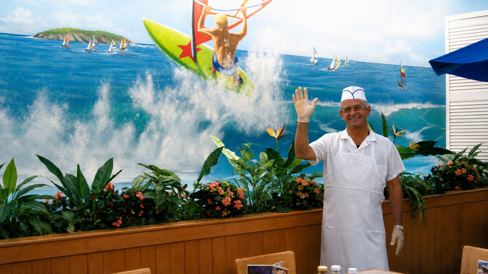 A smiling Surf Buffet employee in a white paper hat, shirt and apron raises a gloved hand in a wave beside the restaurant's painted ocean mural of a windsurfer breaking through a wave.