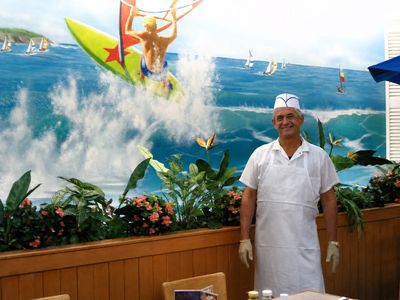 Smiling Surf Buffet employee in a white paper hat and apron standing in front of the painted wave mural, food-service gloves on both hands.