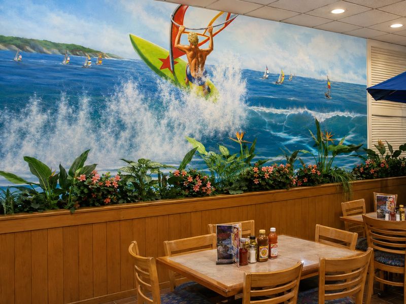 Close view of the Surf Buffet windsurfer mural above wood-panelled wainscot, planter flowers and a dining table set with menus and hot-sauce bottles.
