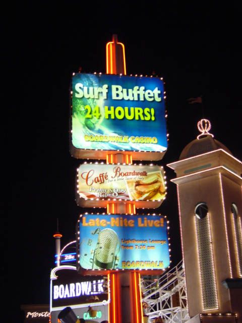 Night photo of a sign pylon with three stacked lit panels: Surf Buffet 24 Hours with wave art, Caffe Boardwalk prime rib, and Late-Nite Live with a chrome microphone graphic, red neon striping the pole.