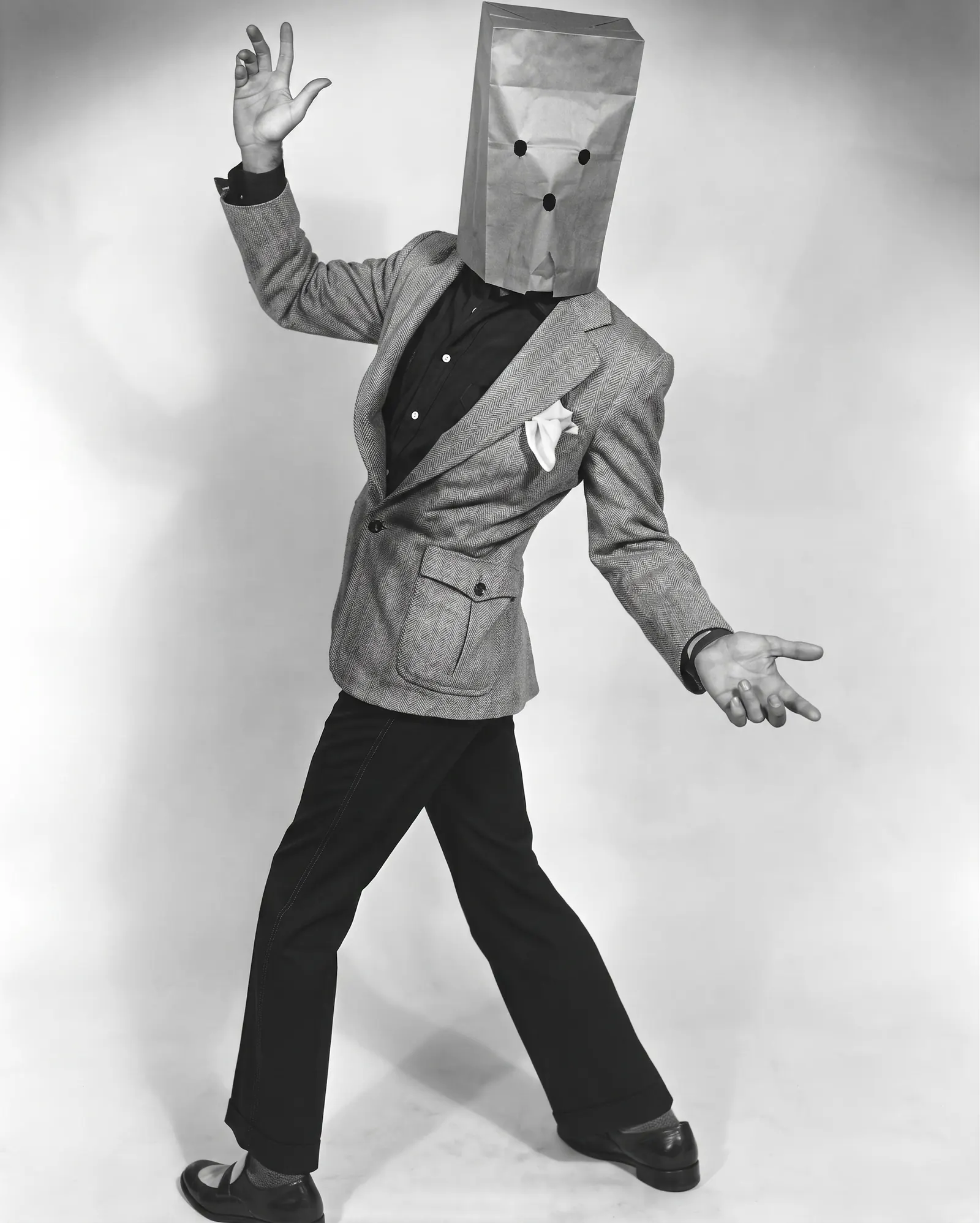 Murray Langston as the Unknown Comic in a grey blazer and paper bag mask, gesturing with both hands against a simple studio background.