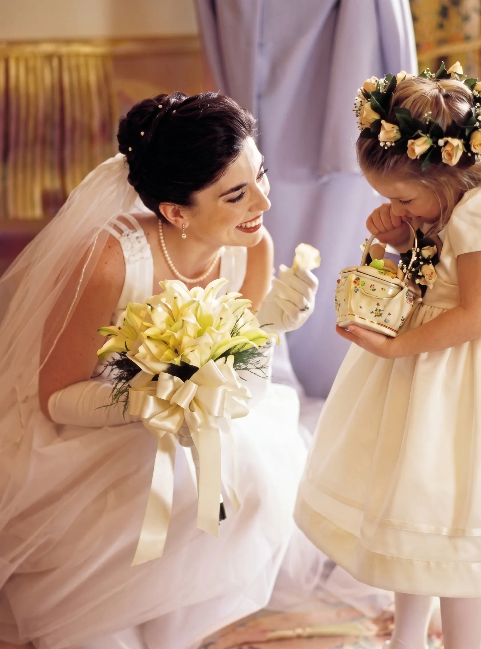 A smiling bride holding white lilies leans toward a flower girl carrying a small floral basket.